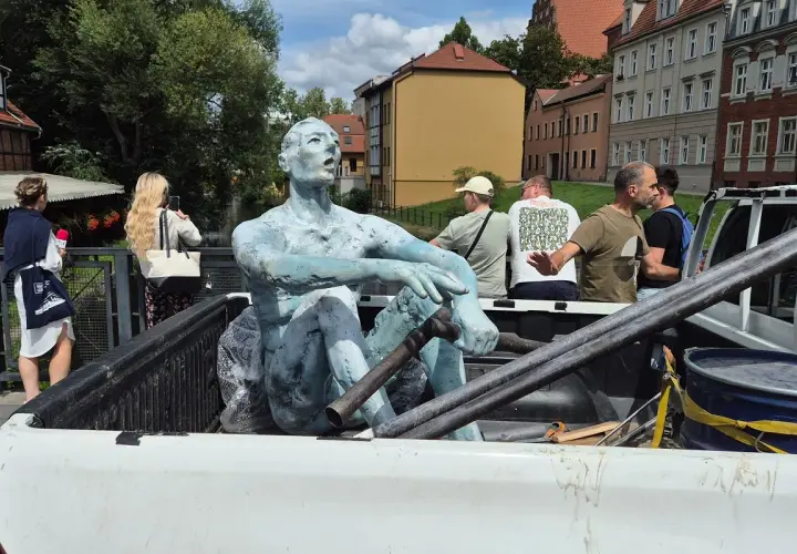 Rzeźbiarze transportują niebieską rzeźbę siedzącego mężczyzny w bagażniku pickupa. W tle widać budynki i ludzi.