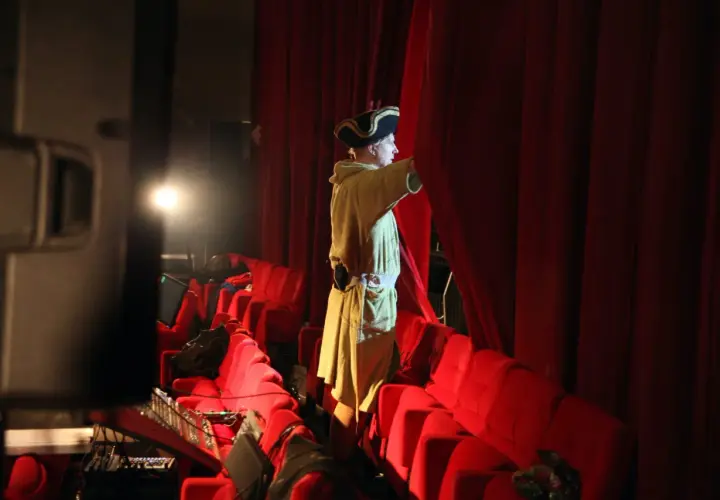 En person i historisk dräkt drar undan en röd ridå på en teater.