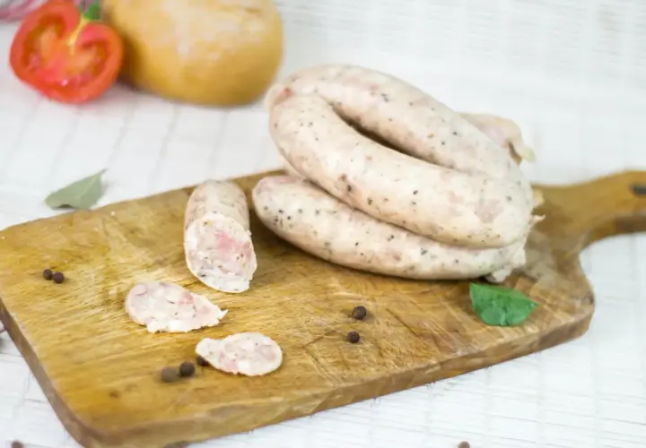 Biała kiełbasa na desce, obok pomidor, czosnek i przyprawy. Zastanawiasz się, ile czasu moczymy jelita do kiełbasy?