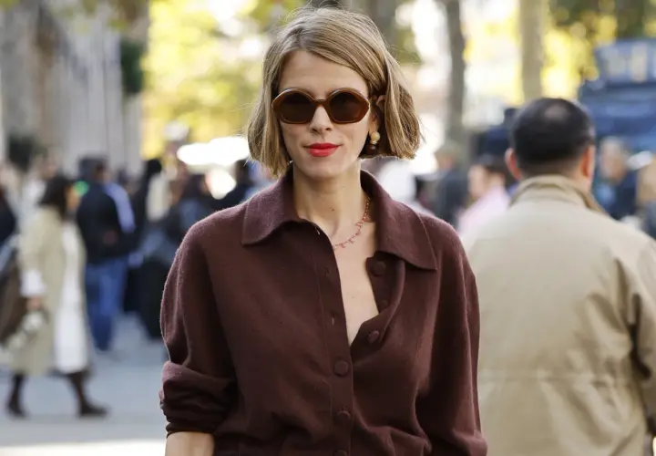 Mujer con corte francés con raya lateral, gafas oscuras y labial rojo, luciendo un suéter marrón.