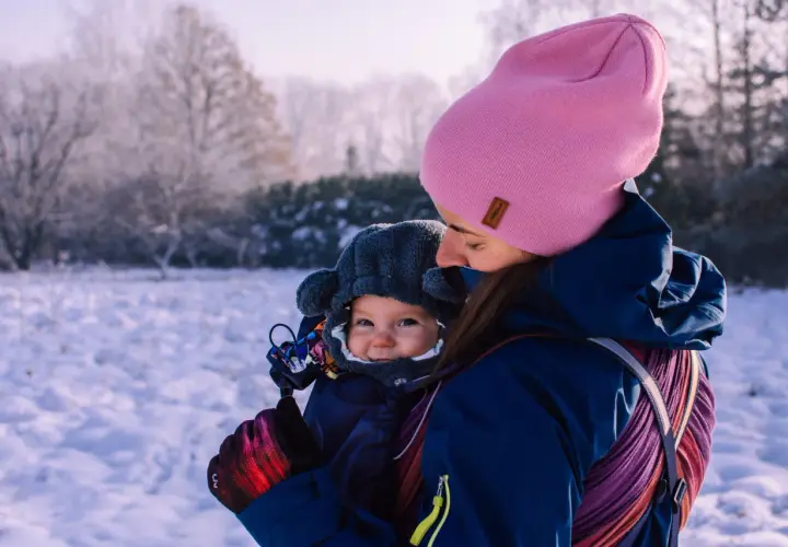 Mama w różowej czapce i granatowej kurtce tuli dziecko w nosidełku. Zima w pełni, śnieg wokół. Jak ubrać dziecko do nosidełka zimą? Warstwowo i ciepło!