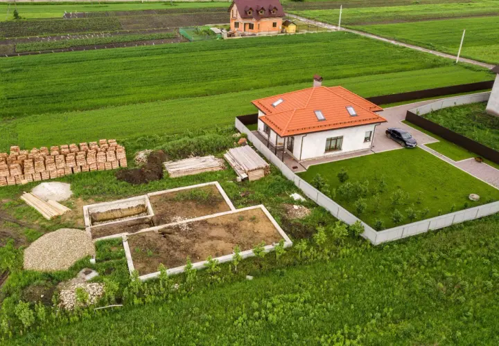 Budowa domu na działce z fundamentami, materiałami budowlanymi i gotowym budynkiem obok. Minimalna powierzchnia działki budowlanej widać na przykładzie.