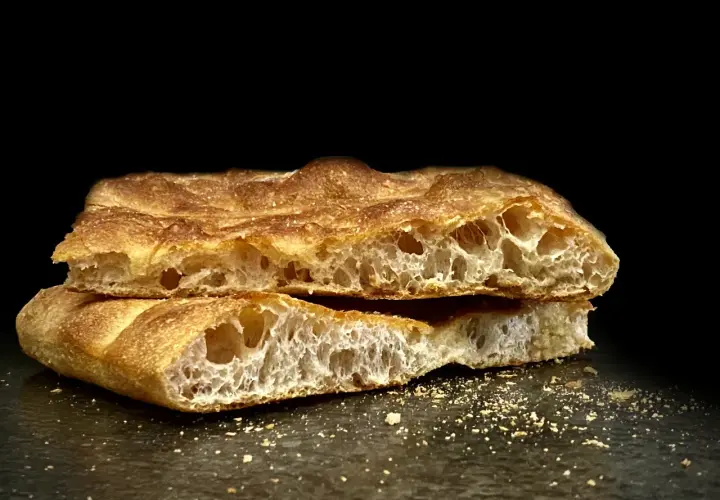 Schiacciata toscana dorata e fragrante, tagliata a metà, con alveolatura evidente e briciole sparse.