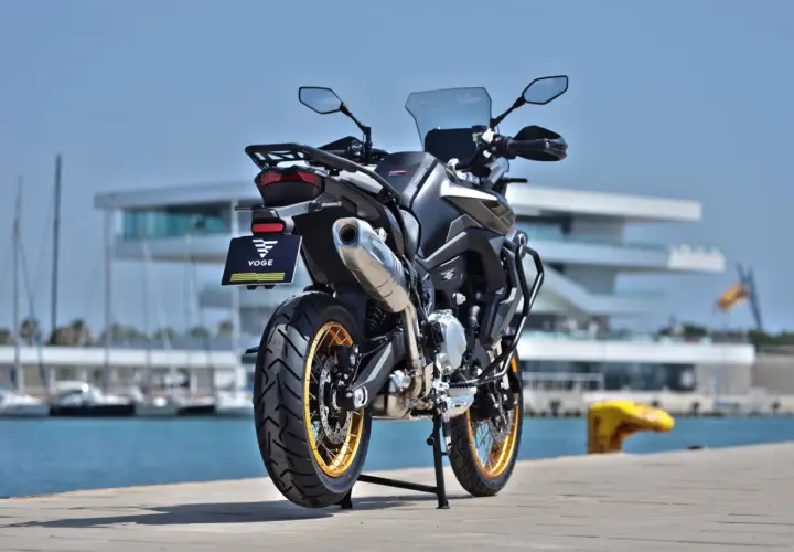 Voge 900 DSX ficha técnica: una moto de aventura negra con detalles dorados, aparcada junto a un puerto con veleros y un edificio moderno.