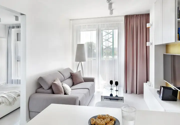 Jasny, bardzo mały salon w bloku z szarą kanapą, stolikiem kawowym i widokiem na balkon. Obok widać łóżko.