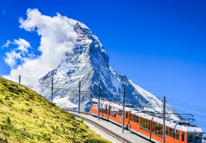 An orange cogwheel train ascends a grassy slope towards the majestic Matterhorn, a perfect scene for hiking in Switzerland.