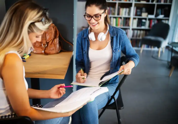 Dwie studentki uczą się do egzaminów w bibliotece. Jedna z nich, w okularach i dżinsowej koszuli, uśmiecha się, pisząc w zeszycie. To idealne miejsce do nauki w **liceum dla dorosłych 2-letnim**.