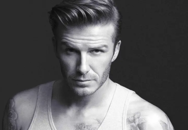Mężczyzna z tatuażami i modną fryzurą side part, w podkoszulku, patrzy intensywnie.