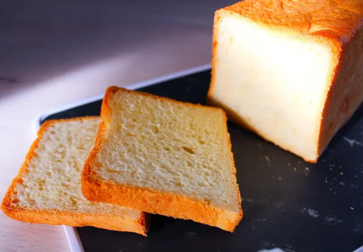 Pain de mie moelleux, parfait pour une recette pain de mie boulangerie maison. Deux tranches dorées et un pain entier sur un plateau sombre.