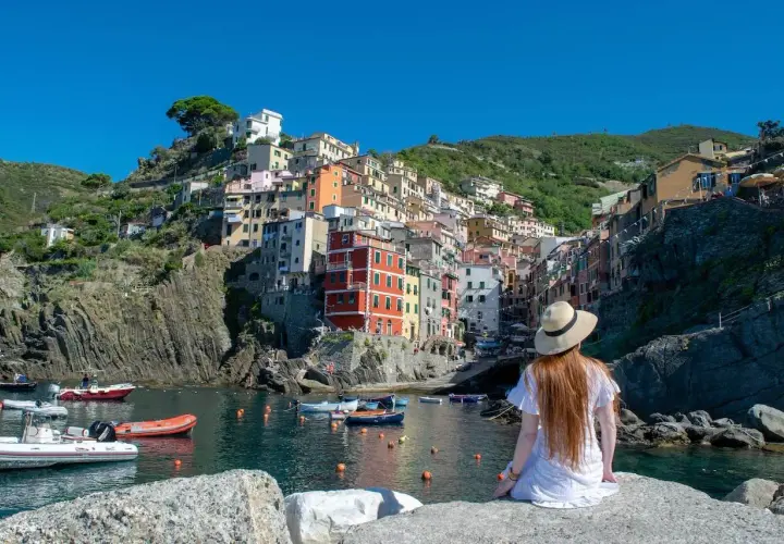 A woman in a white dress and hat gazes at the colorful houses of Riomaggiore, one of the best towns to stay in Cinque Terre, nestled on a cliffside overlooking the harbor.