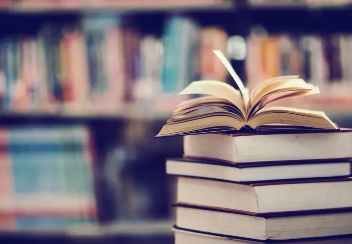 Stos książek, na wierzchu otwarta księga. W tle regały pełne literatury, w tym podręczniki do prawa pracy.
