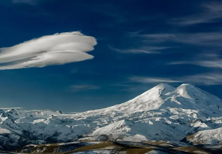 Pokryty śniegiem Elbrus, najwyższy szczyt Rosji, góruje nad ośnieżonymi pasmami górskimi pod błękitnym niebem z charakterystyczną chmurą.