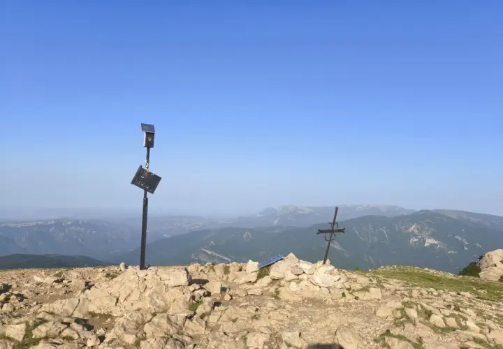 En el cap de la gallina pelada, un poste con placa y un sol solar vigilan el paisaje montañoso. A su lado, una cruz metálica se alza sobre rocas.