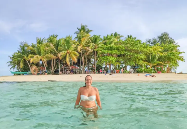 Mujer sonríe en aguas cristalinas, con una isla tropical llena de palmeras al fondo. ¡Un sueño para viajar a Filipinas!