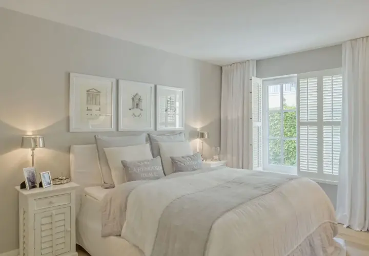 Helles, luftiges Schlafzimmer im Strandhaus-Stil mit weißem Bett, Kissen und Gardinen. Drei gerahmte Zeichnungen über dem Bett.