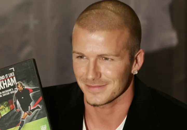 David Beckham, con su característico corte afeitado, sonríe mientras sostiene un DVD de sus habilidades de fútbol.