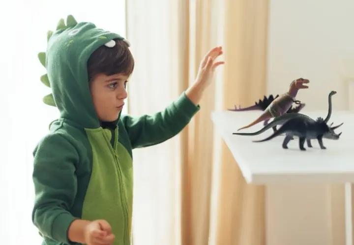 Chłopiec w zielonym stroju dinozaura DIY sięga po figurki prehistorycznych gadów.