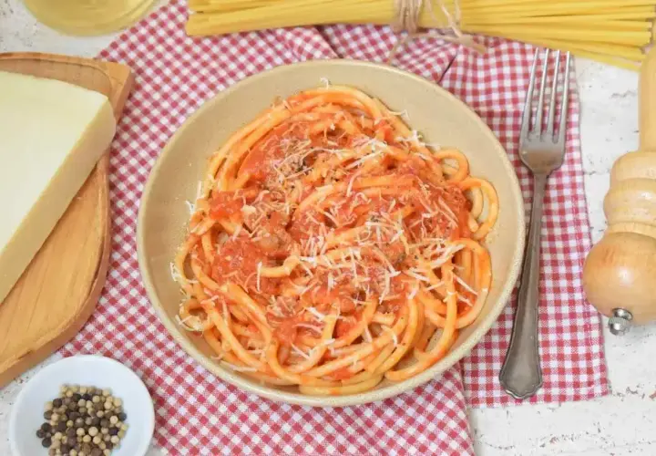 Miseczka pełna aromatycznego makaronu amatriciana, posypanego startym parmezanem. Obok kawałek sera, ziarna pieprzu i widelec.