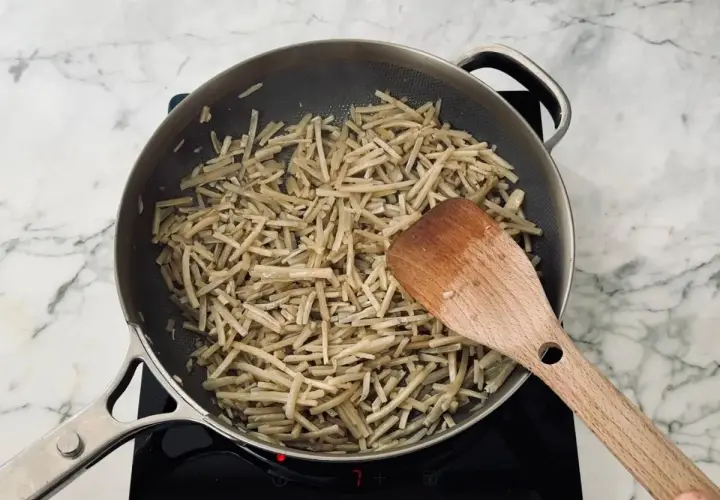 Strips of burdock root are being stir-fried in a pan, a key step in preparing a delicious kinpira recipe. A wooden spatula is used to toss the ingredients.