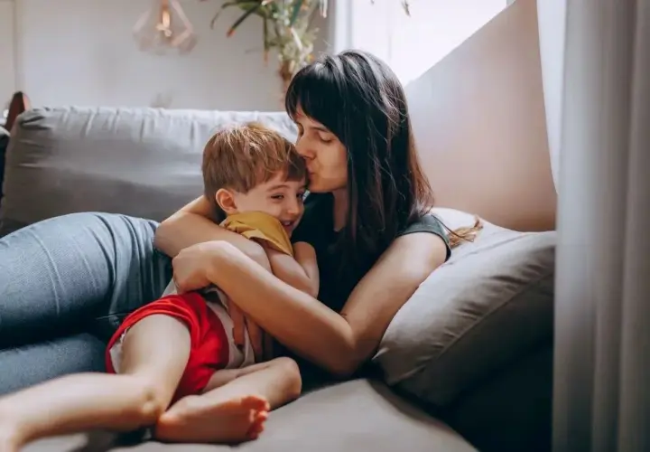 Mama całuje synka w czoło, przytulając go na kanapie. Dziecko śmieje się radośnie. Może dziś nie posyłam dziecka do przedszkola.
