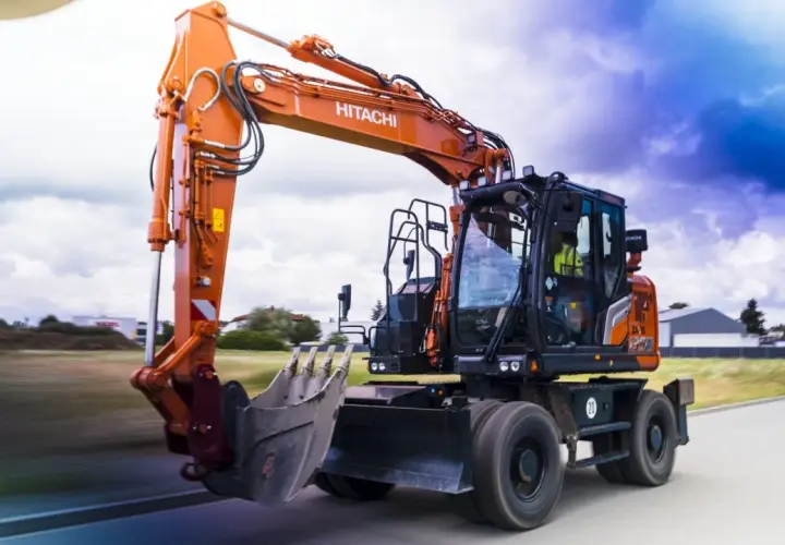 Pomarańczowa koparka Hitachi, gotowa do pracy na złomowisku, pędzi po drodze.