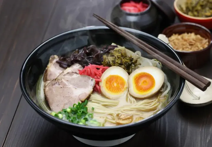 Pyszny ramen co to? Miska parującego bulionu z makaronem, plastrami wieprzowiny, jajkiem na pół, szczypiorkiem i marynowanymi dodatkami.
