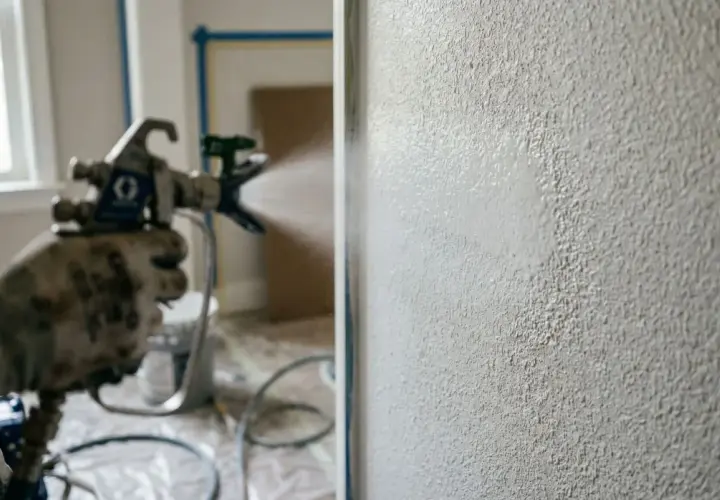 Mano guantata con pistola a spruzzo per imbiancare a spruzzo una parete ruvida.