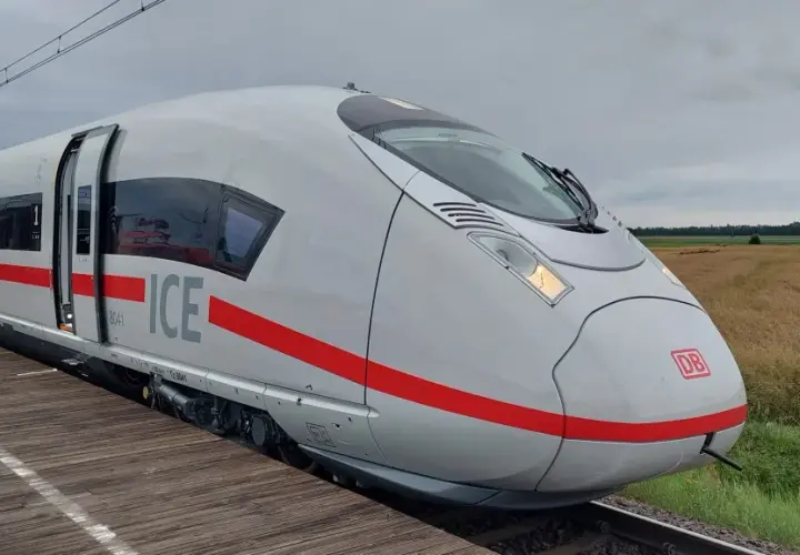 Biały pociąg Siemens Velaro ICE z czerwonym pasem stoi na drewnianym peronie. W tle pole i zachmurzone niebo.