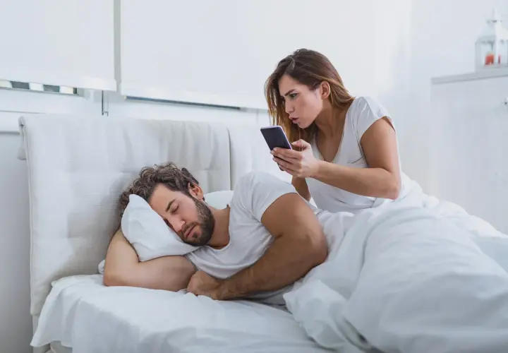 Donna preoccupata controlla il telefono del partner addormentato, un classico esempio di psicologia della sfiducia.