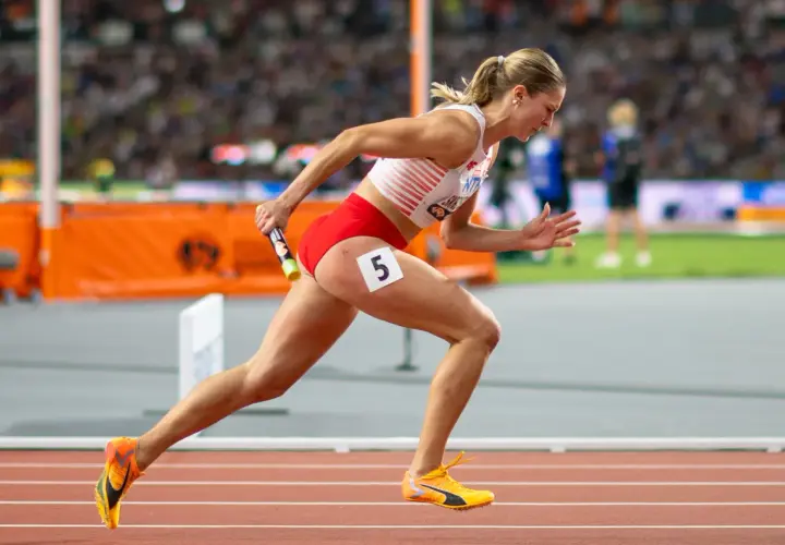 Zawodniczka biegnie z pałeczką w sztafecie 4x100 kobiet. Numer 5 na udzie.