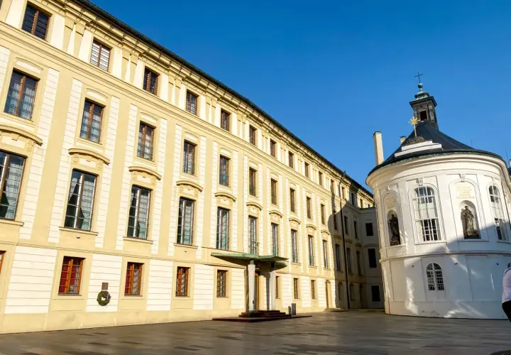 Hradczany zamek w Pradze. Długi, żółty budynek z wieloma oknami i biała, okrągła kaplica pod błękitnym niebem.