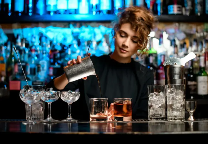 Kto robi drinki? Młoda barmanka z kręconymi włosami przelewa płyn z shakera do szklanki z lodem.