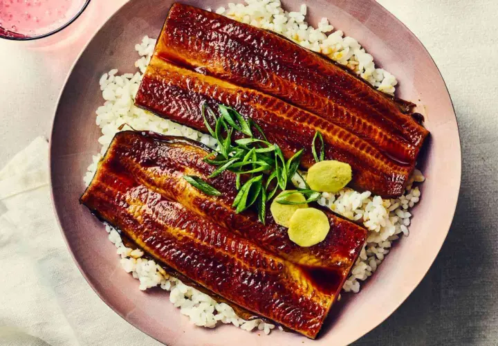 Two glazed unadon fillets rest on a bed of white rice, garnished with green onions and pickled ginger.