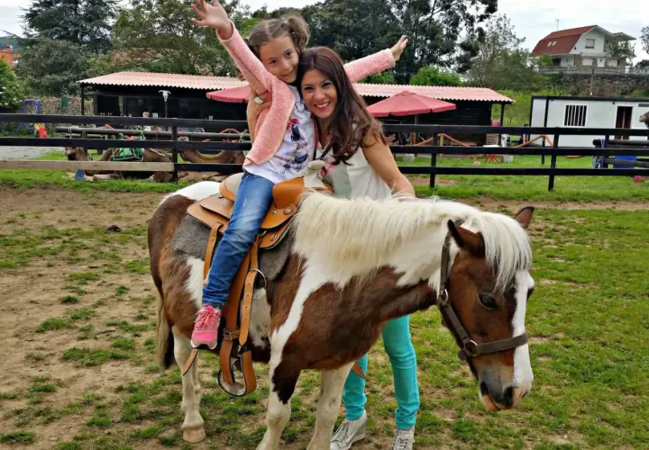 Niña feliz en un poni, disfrutando de una ruta a caballo en Cantabria. Madre sonriente la acompaña.