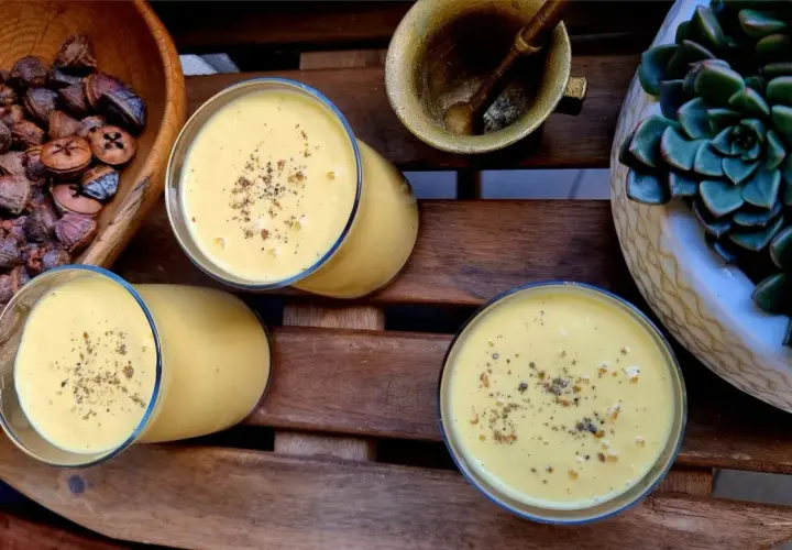 Trzy szklanki kremowego mango lassi, posypane przyprawami, obok miski z suszonymi owocami i sukulentu.