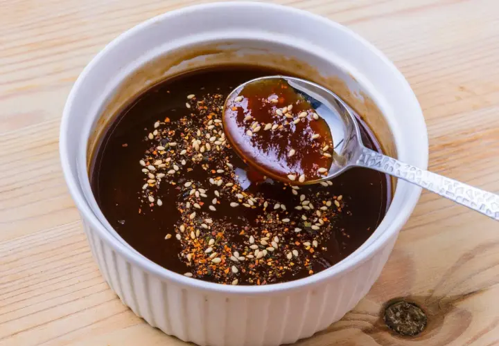 A spoon scoops rich, dark okonomiyaki sauce from a white bowl, sprinkled with sesame seeds and chili flakes. This is how to make okonomiyaki sauce.
