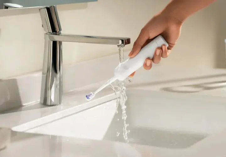 Mano che sciacqua uno spazzolino elettrico sotto un rubinetto. Ideale per chi soffre di gengive ritirate.