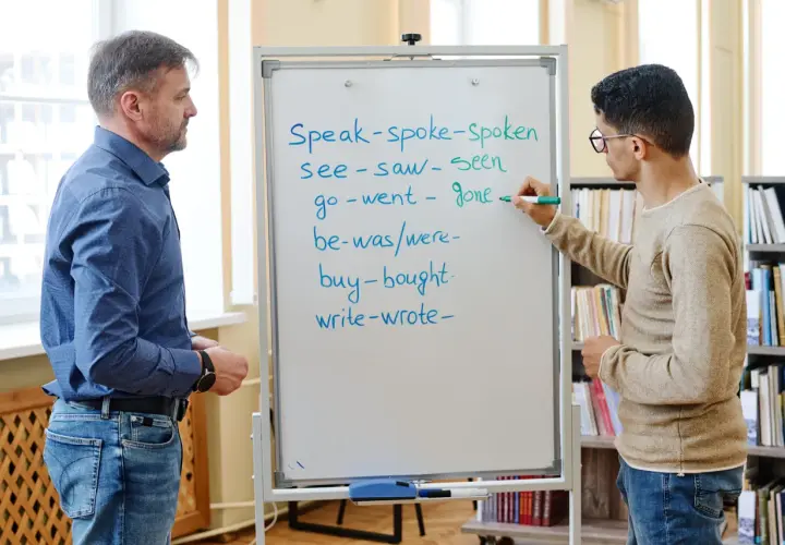 Un insegnante e uno studente studiano il passato in inglese. Lo studente scrive verbi irregolari sulla lavagna.