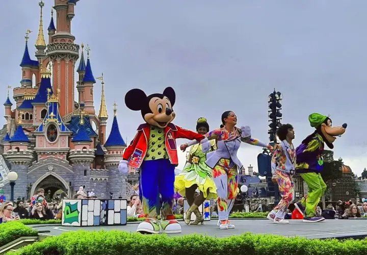 Myszka Miki i Goofy na scenie przed zamkiem w Disneyland Paris, otoczeni tancerzami w kolorowych strojach. Niezapomniane atrakcje Disneyland Paris!