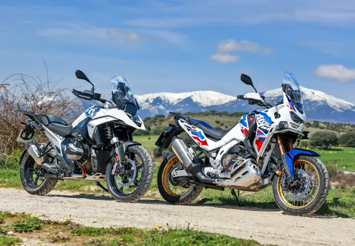 Dos motos de aventura, una Honda Africa Twin 2026 y otra blanca, aparcadas en un camino rural con montañas nevadas al fondo.