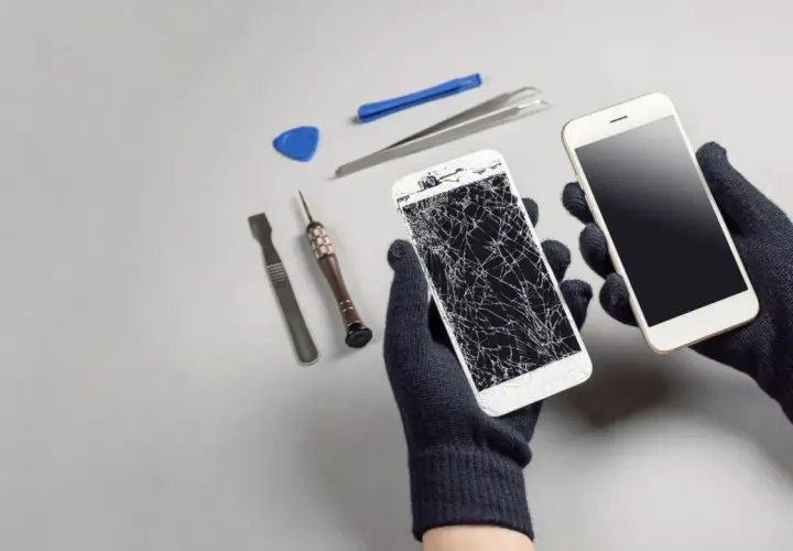 Ręce w rękawiczkach trzymają dwa telefony: jeden z pękniętym ekranem, drugi sprawny. Obok narzędzia do naprawy. Upadek telefonu skutki widoczne.