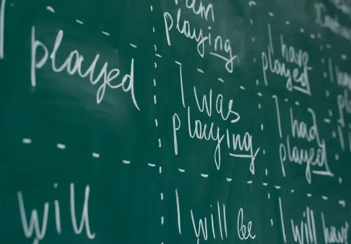 Scrittura a mano su lavagna verde: "I played", "I was playing", "I have played", "I will play". Esercizi di grammatica inglese, con focus sui tempi verbali.