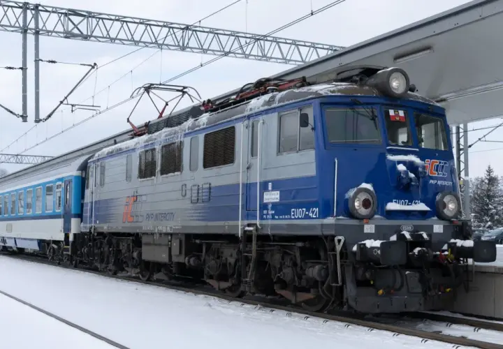 Lokomotywa EU07-421 PKP Intercity z pociągiem na tle zimowego krajobrazu. Widać elementy sieci trakcyjnej i peron.
