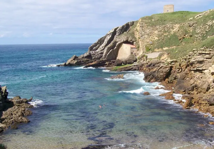 Costa rocosa y mar turquesa en Santillana del Mar, Santander. Una pequeña ermita se esconde en la roca y una torre vigía corona la colina.