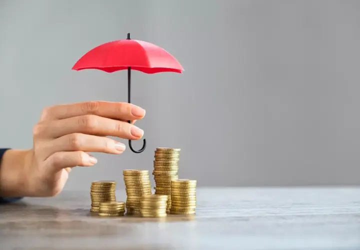 Une main protège des piles de pièces avec un petit parapluie rouge, symbolisant l'assurance PTZ.