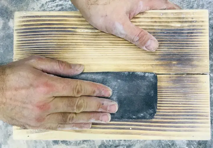 Mani che levigano un pezzo di legno con carta vetrata per sverniciare legno, rimuovendo la vernice vecchia.