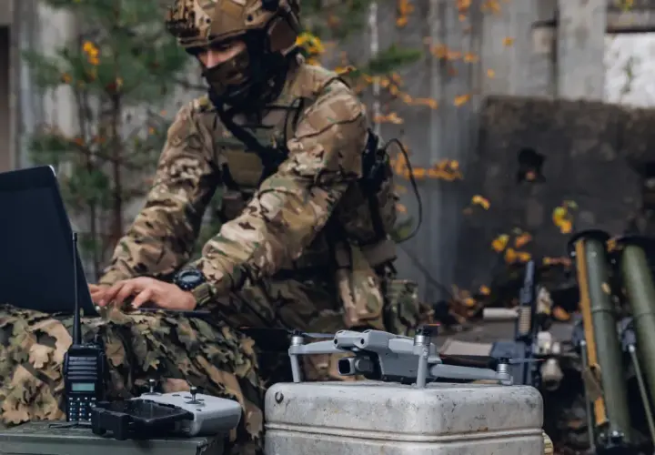 Żołnierz w kamuflażu pracuje na laptopie podczas ćwiczeń wojskowych. Obok leży dron i radiostacja.