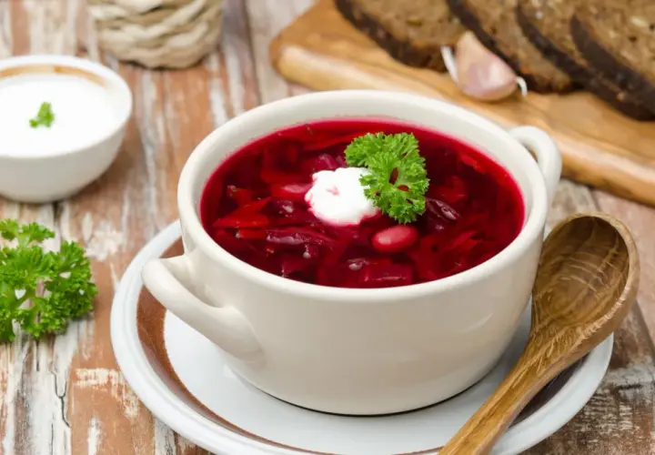Pyszny botwinka przepis na rozgrzewającą zupę z buraków, podana z kleksem śmietany i natką pietruszki. Obok kromki chleba i czosnek.