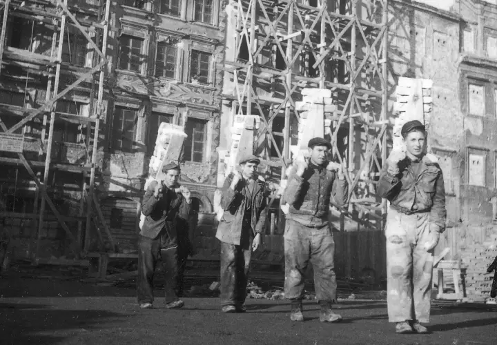Mężczyźni niosą cegły na tle odbudowywanej kamienicy. To obraz Warszawy po wojnie, gdzie trwa mozolna praca nad przywróceniem miasta do życia.