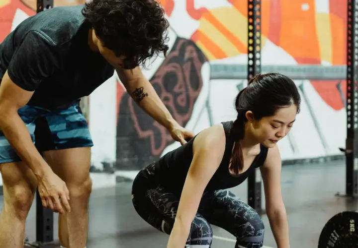 Entrenador ayuda a mujer a levantar pesas en gimnasio con mural colorido de fondo.